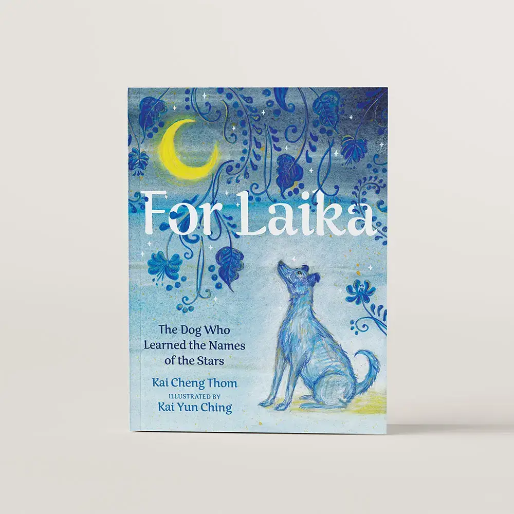 Book cover for the kids book For Laika. The cover is drawn in all blue tones with a dog looking up at the moon. There's ornamental designs of blue floral patters across the top of the cover. 