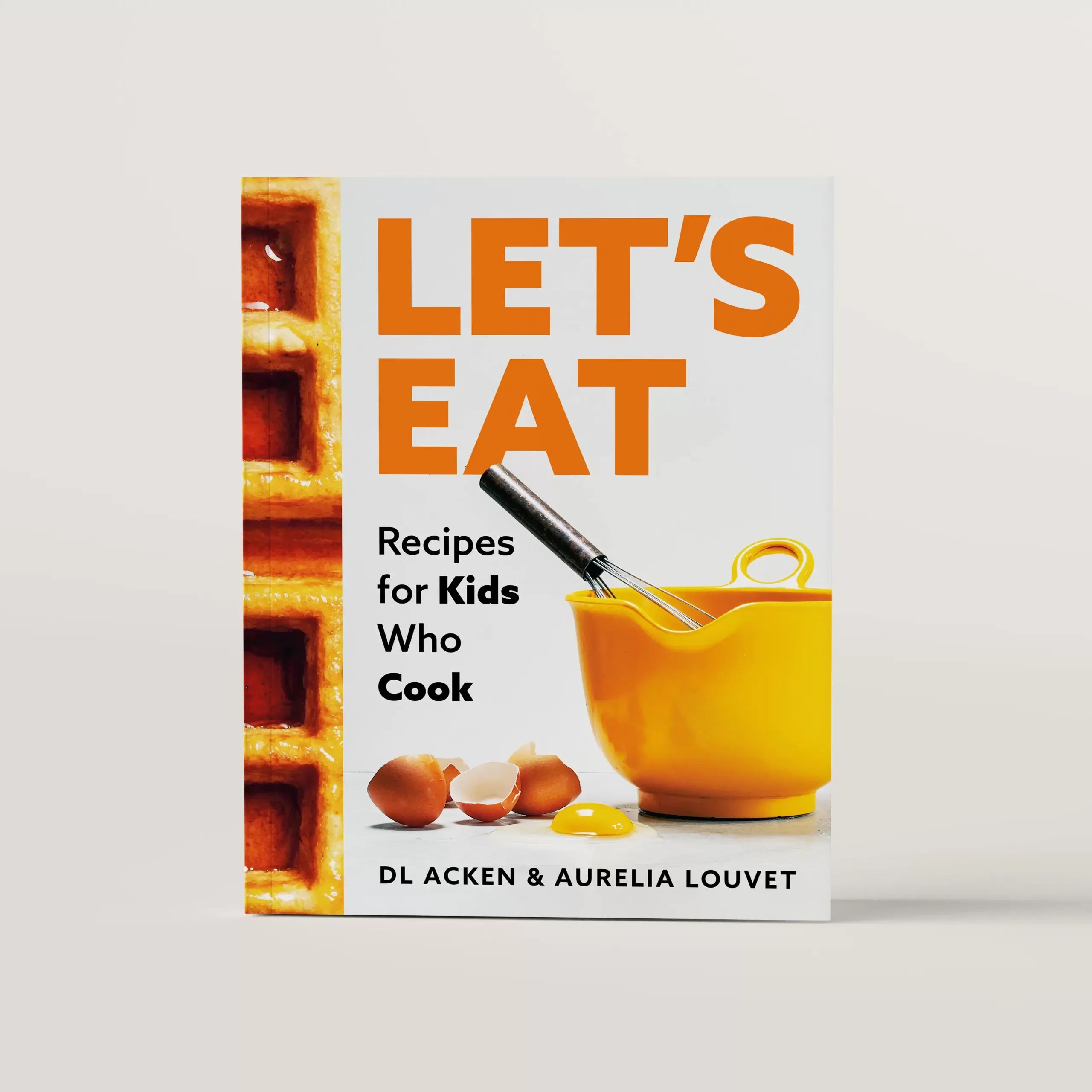 Let's Eat cookbook cover featuring a waffle photo that wraps around the spine. The main photo is of a bright yellow bowl with a whisk and cracked eggs beside it.