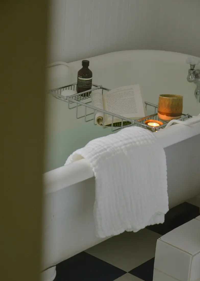 Photo peaking into a bathroom with a claw tub, with a bath drawn, a candle and a book in a tub caddie with a white towel hanging on the edge 