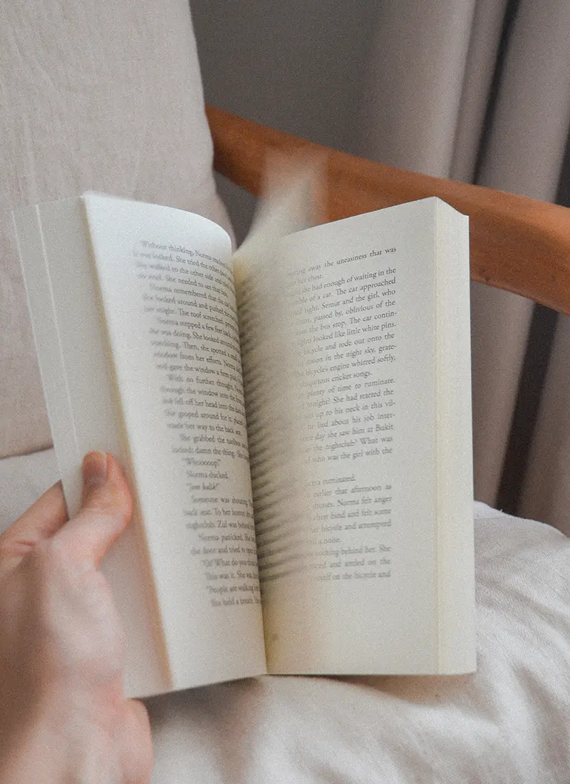 Photo of a book getting flipped, the pages are blurred, the book is held in someones left hand and is resting on a chair